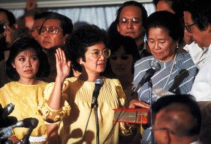 Corazon Aquino, standing amongst a crowd of spectators, being sworn in as President of the Philippines while wearing all yellow including yellow eyeglasses.